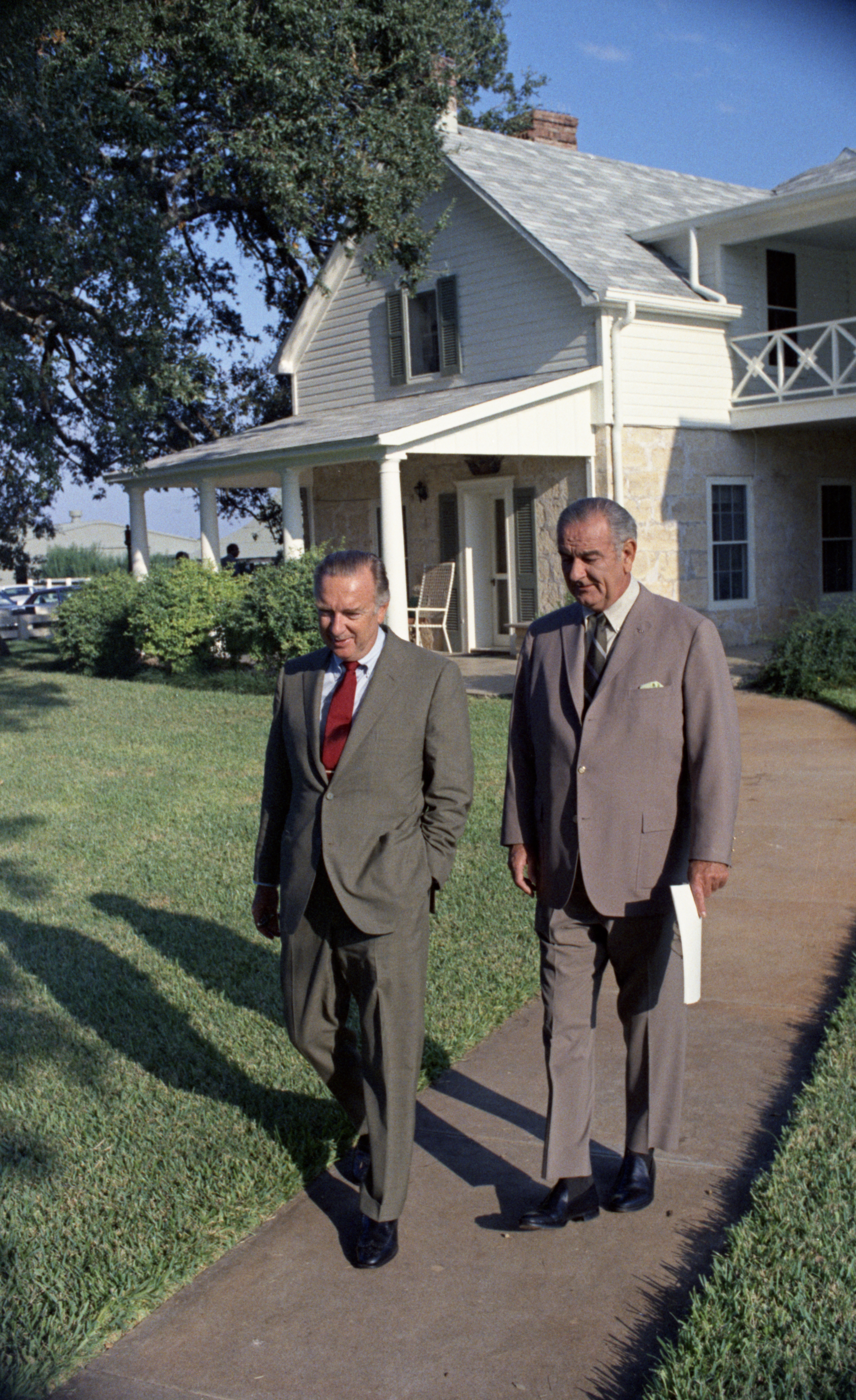 Walter Kronkite and Lyndon Baines Johnson walk down a sidewalk outside a country house. 